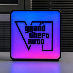 Op de afbeelding: Een vierkante LED-lichtbak met een zwarte rand en een oplichtend oppervlak met een kleurverloop van blauw aan de bovenkant naar roze aan de onderkant. De zwarte tekst Grand Theft Auto VI staat in het midden over een grote Romeinse VI.