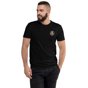 May include: Black short-sleeved t-shirt with a small, detailed emblem on the upper left chest. The emblem features a shield shape with an anchor design and ornate detailing. The shirt is a solid colour and has a classic crew neck.
