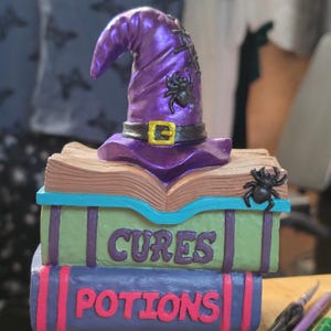 May include: A decorative sculpture featuring a stack of two books topped with an open book and a purple witch hat. The green book is labeled CURES and the purple book is labeled POTIONS. Black spiders are attached to the hat and the open book.