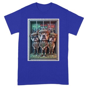 Op de afbeelding: Koningsblauw T-shirt met een afbeelding van drie bulldog-personages in golfkleding. De bulldogs dragen tweed pakken, hoeden en houden golfclubs vast. De tekst "HOLE IN ONE" is inbegrepen.
