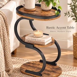 May include: A three-tier accent table with a black metal frame and oval wood-grain shelves. The top shelf holds a mug and a potted plant, the middle shelf holds a candle and two books titled The Kinfolk Home and At Home, and the base is oval.