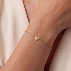 May include: A gold-coloured bracelet with a paw print charm. The charm is the focal point of a fine chain, worn on a wrist. The background is a neutral beige colour.