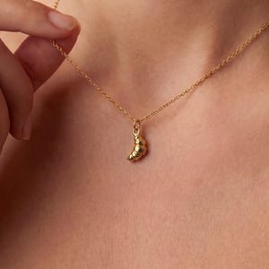 Puede incluir: Un delicado collar de oro con un colgante de croissant. El colgante es una miniatura detallada de un croissant. El collar tiene una cadena fina, y el diseño es elegante y minimalista. Un accesorio de moda.