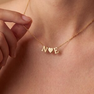 May include: A delicate gold necklace featuring the letters "N" and "E" separated by a heart-shaped charm. The necklace is displayed on a person's neck, showcasing its fine chain and personalized design. The pendant is a stylish accessory.