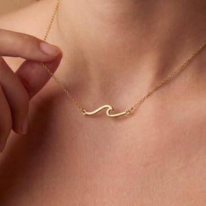 May include: A delicate gold-colored necklace featuring a wave design. The pendant is a simple, curved line, creating a minimalist ocean-inspired aesthetic. The necklace is worn around the neck, showcasing its elegant and subtle design.