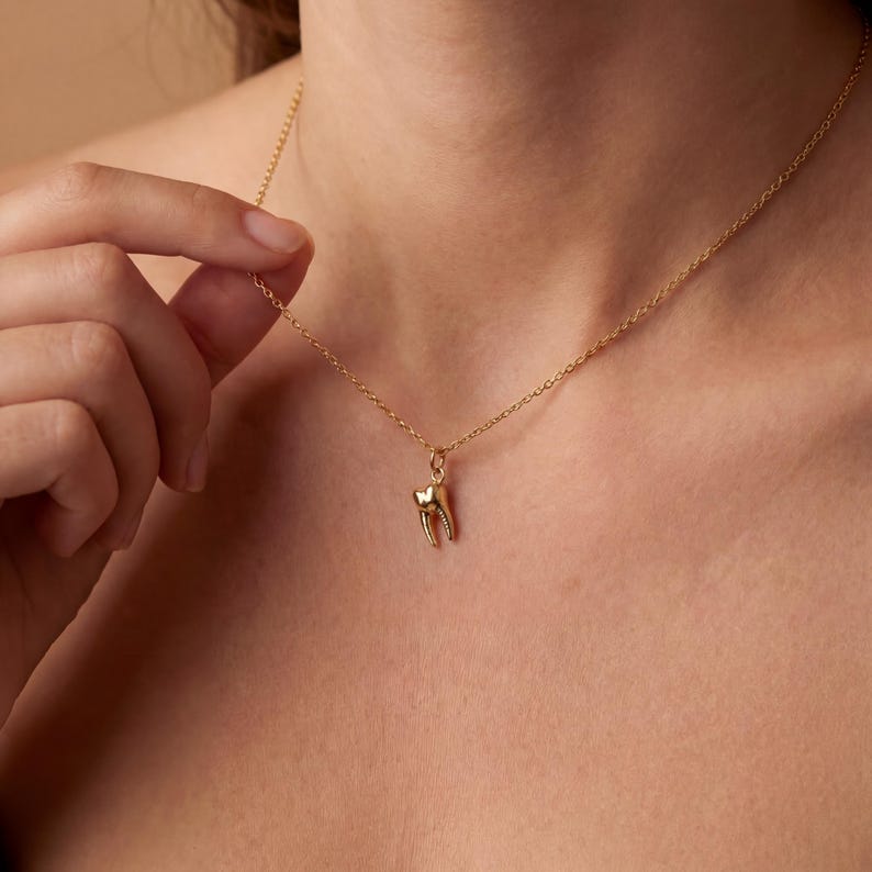 K&ouml;nnte beinhalten: Eine zarte goldene Halskette mit einem kleinen Zahnanh&auml;nger. Der Anh&auml;nger ist eine detaillierte Darstellung eines Molaren, mit sichtbaren Wurzeln und Zahnschmelz. Die feine Kette sorgt f&uuml;r einen minimalistischen und stilvollen Look.