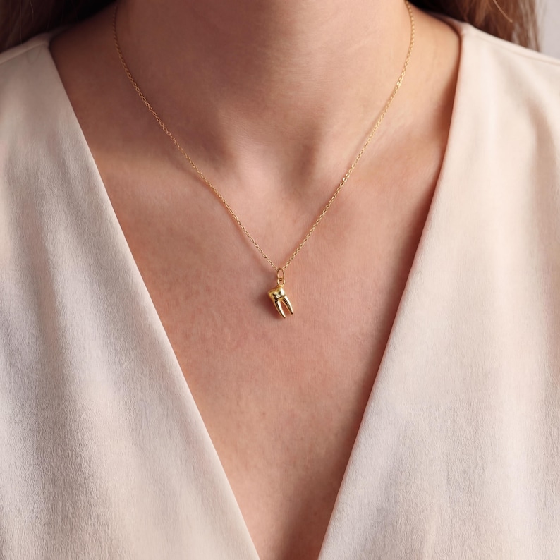 K&ouml;nnte beinhalten: Eine goldfarbene Halskette mit einem kleinen Zahnanh&auml;nger. Der Anh&auml;nger ist eine detaillierte Miniatur eines Backenzahns. Die Halskette ist filigran und scheint aus Gold zu sein. Der Hintergrund ist eine weiche, neutrale Farbe.