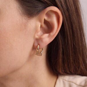 May include: Close-up of a gold hoop earring with a small, rectangular charm. The charm features a layered, linear design. The earring is worn on an ear, with the focus on the jewellery.