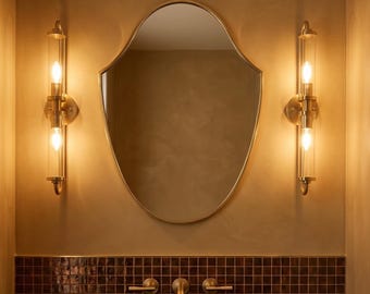May include: A bathroom vanity featuring a dark stone countertop, a rectangular sink, and a fluted wooden cabinet. Above, a shield-shaped mirror is flanked by two vertical brass wall sconces with glass tubes, set against a brown tiled backsplash.