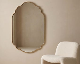 May include: A gold-framed, scalloped-edge mirror hangs on a neutral wall. A cream-coloured, curved armchair sits to the right. The mirror's shape and the chair's design create a modern, minimalist aesthetic.