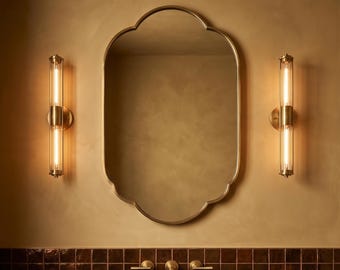 Puede incluir: Interior de baño con espejo dorado, dos apliques de pared con tubos de luz verticales y encimera oscura con lavabo y grifo. La pared está pintada en un tono cálido y neutro, creando un ambiente moderno y elegante.