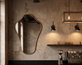 May include: An irregular, organic-shaped wall mirror with a thin gold frame mounted on a textured, weathered concrete wall above a dark wooden counter, featuring a coffee machine, two wooden stools, and a rectangular metal cage pendant light.