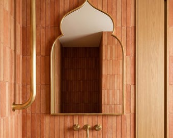May include: A bathroom featuring terracotta-coloured tiled walls and a gold-framed mirror. The marble sink has gold fixtures. A wooden cabinet is on the right, and a gold pipe runs along the wall.