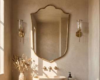 May include: A bathroom vanity featuring a shield-shaped gold mirror, a stone sink, and a wooden cabinet. The vanity is lit by two sconces and natural light. A vase of dried flowers and a bottle of soap are on the countertop.