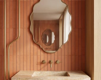 May include: A bathroom with a peach-coloured tiled wall, a gold-framed, scalloped-edge mirror, and a stone sink with gold fixtures. A towel hangs below the sink. The design is modern and minimalist.