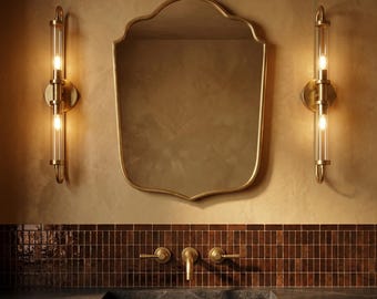May include: A bathroom vanity featuring a shield-shaped mirror, two brass sconces with clear glass tubes, and a dark countertop. The wall is a warm beige, and the backsplash is made of small brown tiles. The vanity has a wooden base.