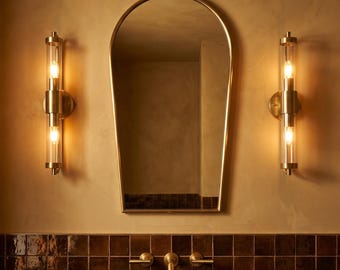 May include: A bathroom interior with a gold-framed mirror, two sconces with cylindrical glass shades, and a dark countertop with a black sink. The wall is a warm, neutral colour, and the fixtures are gold.