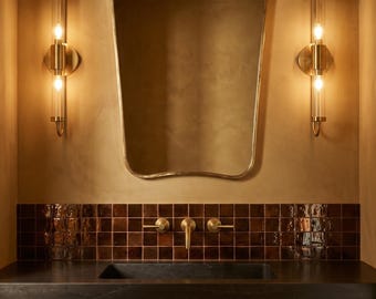 May include: A bathroom vanity area featuring a dark stone countertop, a rectangular sink, and a brass tap. Above, a wavy-edged mirror is flanked by two vertical brass wall sconces with glass tubes. The wall has brown square tiles.