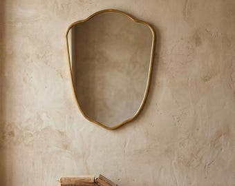 May include: A decorative mirror with a gold frame and an unusual, shield-like outline hangs on a textured beige wall. Beneath it, a wooden bench with rounded legs holds several books. The scene is illuminated by natural light, creating a warm atmosphere.