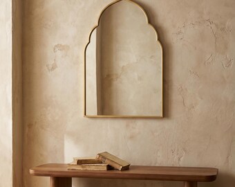 May include: A gold-framed mirror with an arched top hangs above a wooden bench. The mirror reflects the room, and the bench holds two old books. The wall has a textured, neutral-toned finish, creating a warm, inviting atmosphere.