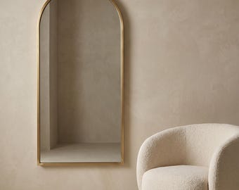 May include: A tall, arched floor mirror with a thin, metallic gold frame. The mirror features a rounded top and straight sides, reflecting a neutral-toned interior space. Beside the mirror sits a rounded, cream-coloured armchair with a textured fabric.
