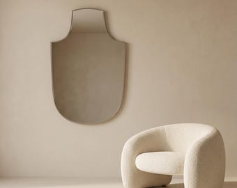 May include: A contemporary interior scene with a shield-shaped mirror, framed in metal, mounted on a beige wall. A cream-coloured, curved armchair is positioned below the mirror, offering a minimalist design.