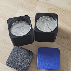 May include: Two black square coin holders containing silver American Eagle coins. One holder is open, revealing the eagle design, while the other shows the Lady Liberty design. Two square lids are placed in front, one black and one blue.