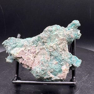 May include: A raw mineral specimen featuring a textured surface with mottled shades of teal, green, and pale pinkish-white. The irregular, jagged rock is displayed on a black metal wire stand, showcasing its natural, unpolished crystalline structure.