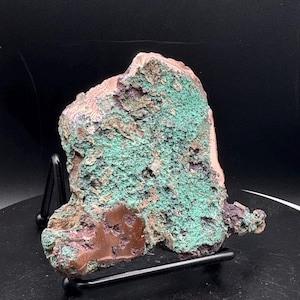 May include: A raw mineral specimen featuring a textured surface with vibrant green malachite deposits, patches of deep reddish-brown copper ore, and dark purple mineral inclusions, displayed on a black metal wire stand against a black background.