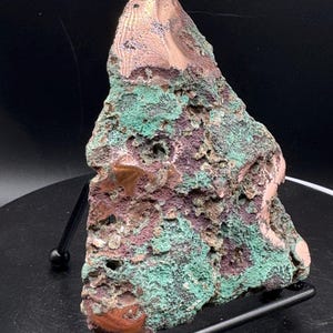 May include: A raw mineral specimen featuring a triangular shape with a rough, porous texture. The surface displays a mix of deep copper-brown, dark purple, and vibrant turquoise-green mineral deposits, resting on a black metal display stand.
