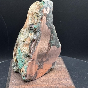 May include: A raw mineral specimen featuring a large, smooth, metallic copper-colored section with a textured surface, contrasted against a rough, jagged matrix of earthy brown, tan, and vibrant green mineral deposits.