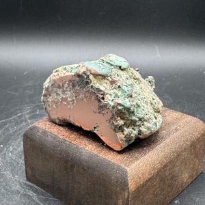 May include: A raw copper specimen featuring a smooth, polished metallic face and a rough, oxidized exterior with green and brown mineral deposits. The specimen is mounted on a dark, rectangular wooden block, showcasing a contrast between the natural, weathered texture and the refined, flat surface of the metal.