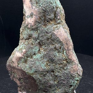 May include: A raw, irregular copper specimen featuring a rough, oxidized surface with prominent patches of green patina and exposed metallic copper areas. The mineral chunk is displayed upright on a simple, thin, silver-toned metal wire stand.