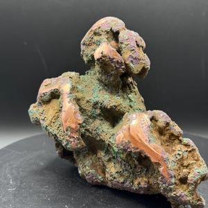May include: A raw copper mineral specimen featuring a rough, porous texture with a mix of metallic copper, oxidised green, and earthy brown tones. The structure is irregular and jagged, showcasing exposed shiny copper veins throughout the surface.