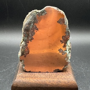 May include: A polished copper specimen with a smooth, metallic orange face and rough, textured edges featuring dark brown and green mineral deposits, mounted upright on a rectangular wooden block base for display purposes.
