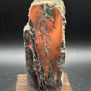 May include: A vertical mineral specimen featuring a polished, smooth, reddish-brown center section with visible grain patterns, flanked by rough, textured, dark grey and green mineral deposits along the edges, mounted on a square wooden base.