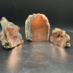 May include: Three raw copper specimens with exposed metallic orange-brown interiors and rough, textured green and brown outer crusts. The pieces vary in size and shape, displaying a distinct contrast between the smooth, polished-looking metallic faces and the jagged, mineralized exterior surfaces.