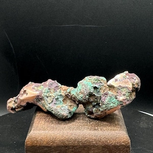May include: A raw, irregular mineral specimen featuring a textured surface with a mix of oxidized copper green, deep purple, and earthy brown tones. The specimen has a porous, craggy appearance with visible metallic copper highlights throughout.