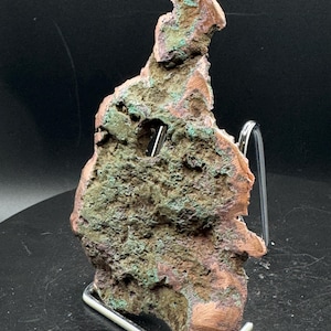May include: A raw copper specimen featuring a rough, textured surface with oxidized green patches and metallic reddish-brown tones. The mineral formation has an irregular, jagged shape with a small natural hole near the top center, displayed upright.