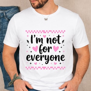Puede incluir: Camiseta blanca con el texto "I'm not for everyone" en letras negras, corazones rosas y un borde a cuadros rosa y blanco. La camiseta tiene mangas cortas.