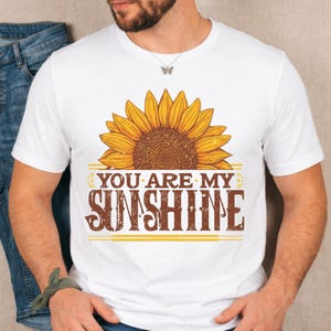 May include: White t-shirt featuring a large sunflower graphic with brown and yellow tones. The text "YOU ARE MY SUNSHINE" is printed below the sunflower in a brown, vintage-style font. The shirt is a casual wear item.