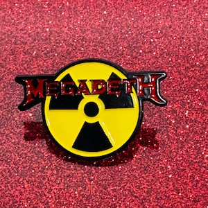 Puede incluir: Un pin de esmalte amarillo y negro con la palabra "Megadeth" en letras rojas. El pin presenta un símbolo nuclear estilizado.
