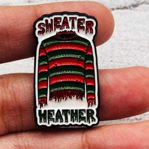 May include: A rectangular pin featuring a red and green striped sweater design with the words "SWEATER" and "HEATHER" in a dripping font. The pin is outlined in black and white.