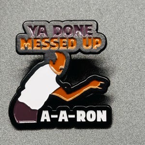 Pode incluir: Um pin de esmalte preto, branco e castanho com uma figura estilizada e o texto "YA DONE MESSED UP" e "A-A-RON". O pin tem um acabamento brilhante e está sobre um fundo cinzento.