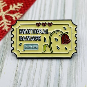 May include: A yellow enamel pin with a ticket design. The pin features a wilted red rose with a green stem and thorns. The text on the pin reads "Emotional Damage book club".