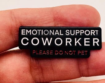 May include: A black rectangular pin with white text that reads "EMOTIONAL SUPPORT COWORKER" and "PLEASE DO NOT PET" in red. The pin is held in a person's hand.