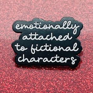 May include: Black enamel pin with white lettering that reads "emotionally attached to fictional characters".