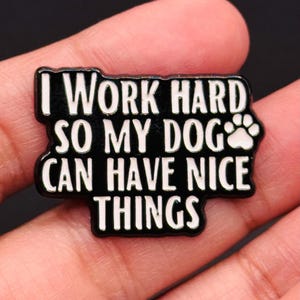 以下が含まれることがあります： 白い文字で「I WORK HARD SO MY DOG CAN HAVE NICE THINGS」と書かれた黒いエナメルピン。ピンには犬の足跡のグラフィックが描かれています。ピンは角が丸い長方形です。