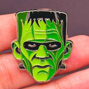 May include: A green and black enamel pin featuring the face of Frankenstein's monster. The pin has a silver border and depicts the monster's iconic features, including black hair, stitched facial details, and a green complexion.