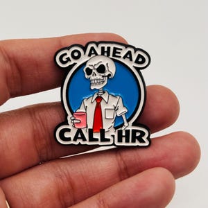 Puede incluir: Un pin de esmalte en blanco y negro con un esqueleto con camisa blanca y corbata roja, sosteniendo una taza rosa. El pin tiene el texto "GO AHEAD CALL HR" en letras blancas sobre un fondo negro.